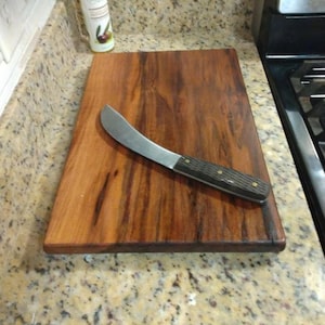 Op de afbeelding: Een rechthoekige houten snijplank met een rijke, donkerbruine houtnerf en een gladde afwerking. Diagonaal op de plank ligt een keukenmes met een gebogen zilverkleurig lemmet en een donker, getextureerd handvat dat is vastgezet met twee koperen klinknagels.