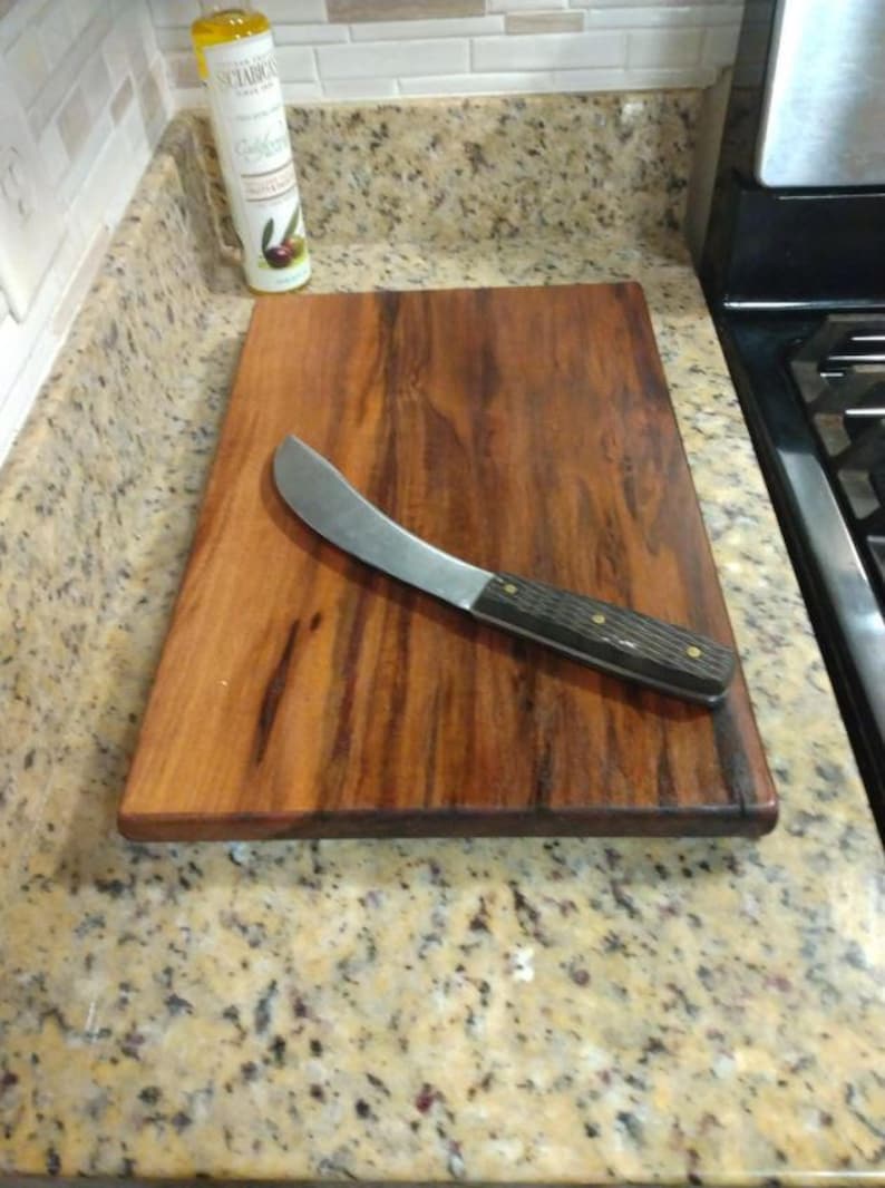 Op de afbeelding: Een rechthoekige houten snijplank met een rijke, donkerbruine houtnerf en een gladde afwerking. Diagonaal op de plank ligt een keukenmes met een gebogen zilverkleurig lemmet en een donker, getextureerd handvat dat is vastgezet met twee koperen klinknagels.
