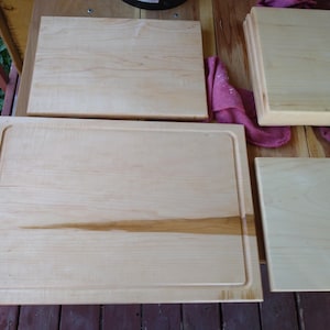 May include: Three rectangular light-coloured wooden cutting boards of varying sizes. The largest board features a recessed juice groove along its perimeter. The wood grain is visible across the smooth, flat surfaces of all three boards.