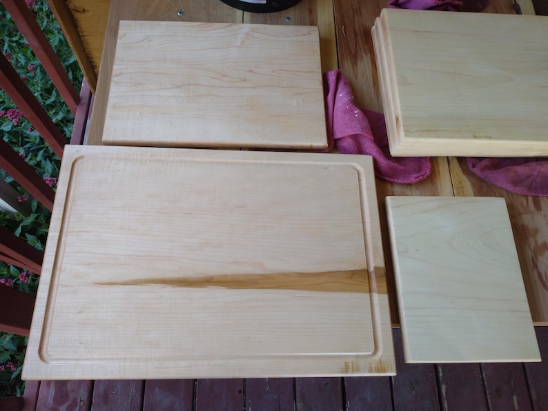 May include: Three rectangular light-coloured wooden cutting boards of varying sizes. The largest board features a recessed juice groove along its perimeter. The wood grain is visible across the smooth, flat surfaces of all three boards.