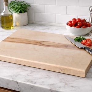 May include: A rectangular wooden cutting board with a light wood grain. A stainless steel knife with a black handle rests on the board, next to a few cut tomatoes and parsley. A bowl of cherry tomatoes is in the background.