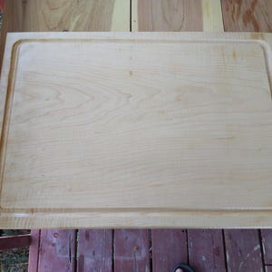 May include: A rectangular light-coloured wooden chopping board featuring a smooth surface with a visible natural grain pattern and a carved juice groove running along the entire perimeter of the board's top face.