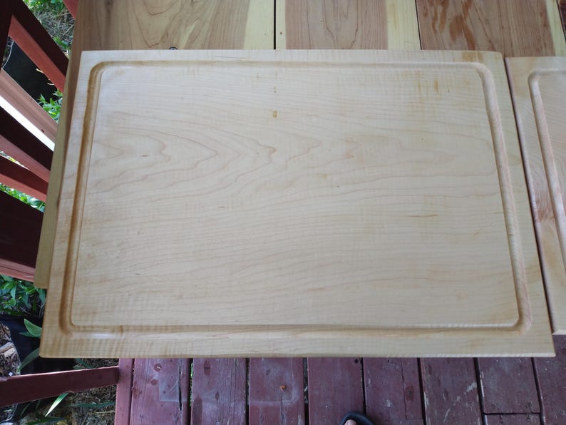 May include: A rectangular light-coloured wooden chopping board featuring a smooth surface with a visible natural grain pattern and a carved juice groove running along the entire perimeter of the board's top face.