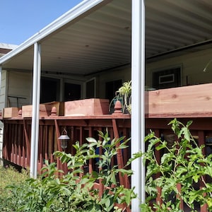 May include: A wooden deck with a red railing supports several rectangular wooden planters. The deck is covered by a white-paneled roof. Green plants and foliage are in the foreground, with a house in the background.