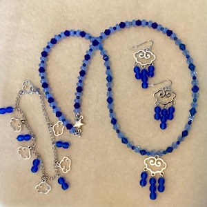 May include: A jewelry set featuring a necklace, bracelet, and earrings. The necklace and bracelet are made of blue beads, with silver cloud charms. The earrings have cloud-shaped charms with dangling blue beads. The necklace has a cloud pendant.