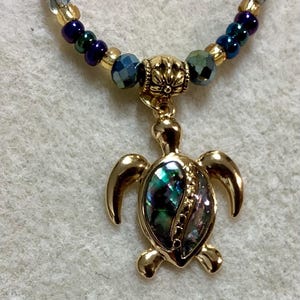 May include: Gold-toned turtle pendant necklace with a colorful abalone shell inlay. The necklace features a beaded strand with gold, blue, and green beads. The pendant is a detailed turtle design.
