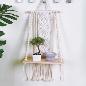 May include: A macrame wall hanging with a wooden shelf, adorned with a small bonsai tree in a white pot and a lavender-colored decorative bowl. The macrame design features diamond patterns and wooden beads.