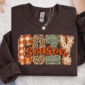 May include: A dark chocolate brown crewneck sweatshirt featuring the word COZY in large, block-style letters with varied patterns including orange plaid, leopard print, sage green, and floral, with the word season in orange script.