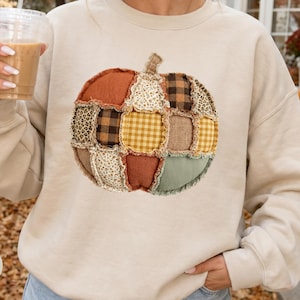 May include: A sand-colored crewneck sweatshirt featuring a large, central graphic of a pumpkin constructed from various fabric patches. The pumpkin design includes patterns such as brown buffalo plaid, leopard print, yellow gingham, and solid orange.