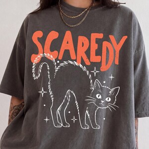 May include: A charcoal gray short-sleeve t-shirt featuring the word SCAREDY in large orange letters above a white line-art illustration of an arched-back cat surrounded by small star shapes. The shirt has a crew neckline and a relaxed fit.