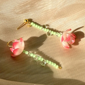 May include: A pair of floral earrings featuring pink and white tulip-shaped pendants. The earrings have a light green beaded stem with clear beads and gold-colored ear wires. The background is a light beige fabric.