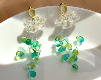 May include: A pair of dangle earrings featuring a floral design with clear, iridescent petals and gold-colored posts. Each earring has a cluster of green and yellow teardrop-shaped beads suspended from delicate strings.