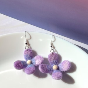 May include: A pair of handmade flower earrings. Each earring features a four-petal design in shades of purple and blue, with a small, cream-colored center. The earrings are displayed on a white plate against a purple and pink background.