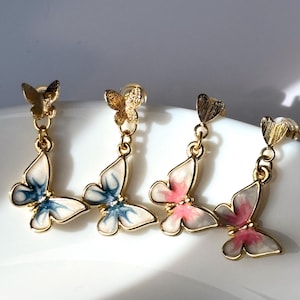 May include: Gold-toned butterfly earrings with dangling butterfly pendants. Two pairs feature white wings with blue accents, and the other two have white wings with pink accents. The earrings are displayed on a white surface.