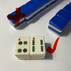 May include: A set of rectangular mahjong tiles with blue and white backs, arranged in rows. Two cream-colored tiles are placed in the foreground, one showing four black and green circular symbols with a red number four, and one showing two green bamboo-like symbols with a red number four.