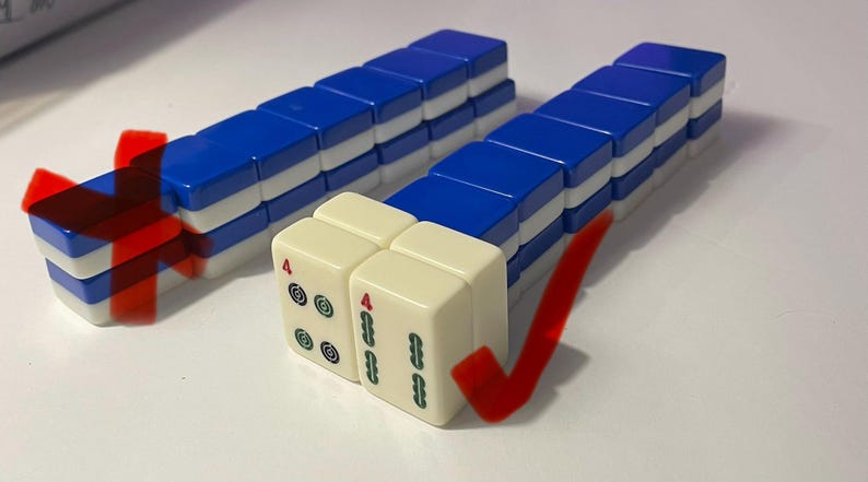 May include: Two rows of blue and white Mahjong tiles arranged in stacks. One row is marked with a red X, and the other row features two cream-colored tiles at the front, each displaying a red number 4 and green symbols, marked with a red checkmark.