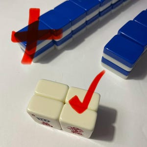 May include: A set of rectangular game tiles with blue backs and white fronts, arranged in rows and stacks. Two off-white tiles are positioned in the foreground, each featuring a small red graphic symbol on the bottom edge of the front face.