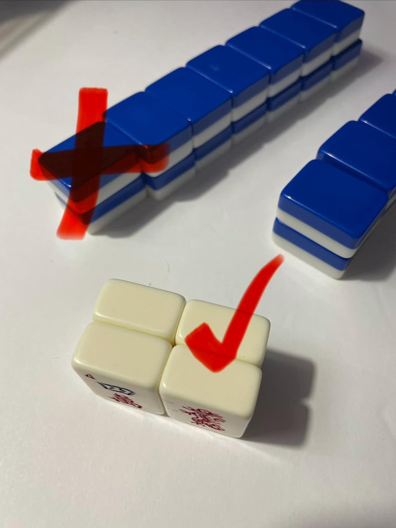 May include: A set of rectangular game tiles with blue backs and white fronts, arranged in rows and stacks. Two off-white tiles are positioned in the foreground, each featuring a small red graphic symbol on the bottom edge of the front face.