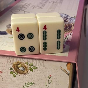 May include: Two off-white rectangular Mahjong tiles standing upright. The left tile features a red number 4 above four circular green and black symbols. The right tile displays a red number 4 above four vertical green bamboo-style symbols.