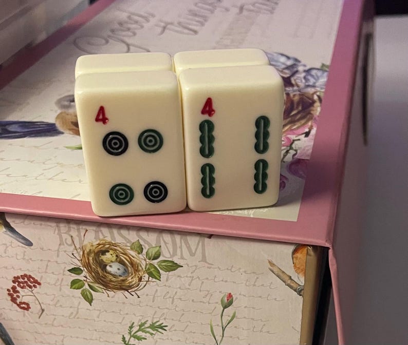 May include: Two off-white rectangular Mahjong tiles standing upright. The left tile features a red number 4 above four circular green and black symbols. The right tile displays a red number 4 above four vertical green bamboo-style symbols.