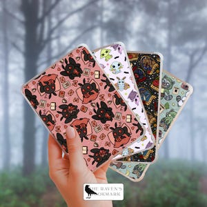 May include: A hand holding four phone cases with unique designs. The cases feature various illustrations, including a black moth, a green creature, a raccoon, and other whimsical creatures. The cases have a clear border and a pink, black, white, and green color scheme.