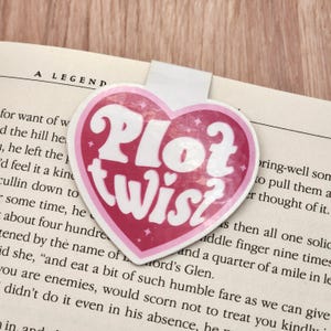 May include: A heart-shaped bookmark with the words "Plot Twist" in white, outlined in pink, and set against a pink background. The bookmark is placed on the pages of a book.