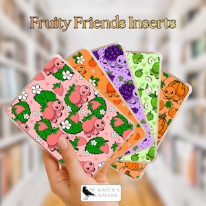 May include: A hand holding five clear phone inserts with colorful fruit and turtle designs. The inserts feature strawberries, oranges, grapes, apples, and pumpkins. The text "Fruity Friends Inserts" is at the top.