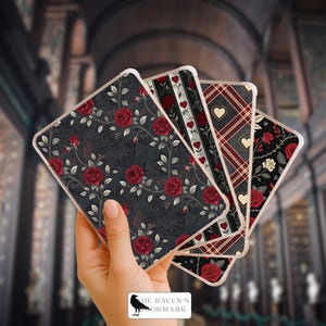 May include: A hand holding four rectangular cards with decorative floral and heart designs. The cards feature red roses, hearts, and plaid patterns on a dark background. The Raven's Bookmark logo is visible.