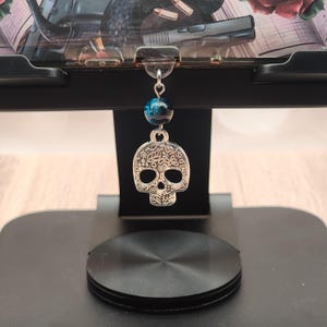 May include: A silver-colored skull charm with intricate floral detailing hangs from a black stand. The charm is suspended by a blue and black bead. The stand appears to be holding a book or tablet with a floral and gun design.