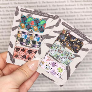 May include: Mini magnetic bookmarks with various colorful designs. One set features cartoon sloths and other creatures, while another showcases a celestial theme with stars and moons. The text "Mini Magnetic Bookmarks" is printed on the cards.