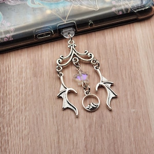 May include: Silver-toned phone charm with a decorative design. The charm features a filigree top, bat-wing shapes, a star-shaped crystal, and a small mountain scene. The charm is attached to a clear phone case.