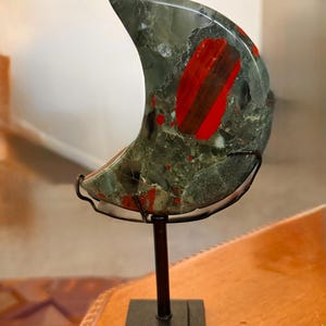May include: A crescent-shaped decorative stone sculpture in shades of green and red, mounted on a black metal stand. The stone has a polished surface with a marbled appearance. The stand has a square base and a vertical support.