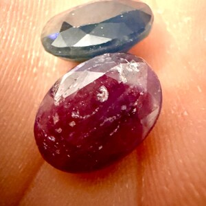 May include: Two oval-cut gemstones. One is a deep blue color, and the other is a rich, dark red. Both stones have a faceted surface, catching the light and displaying their internal structure. The gems are likely for jewelry making.