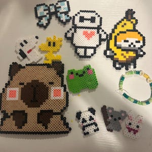 May include: A collection of colorful Perler bead creations, including a bow, a robot with a heart, a banana character, a pug, a frog, a panda, a koala, a bunny, Snoopy, and Woodstock. A beaded bracelet is also included.