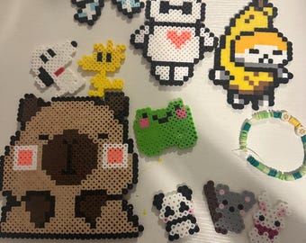 Op de afbeelding: Een verzameling kleurrijke strijkkraalcreaties, waaronder een strik, een robot met een hart, een bananenpersonage, een mopshond, een kikker, een panda, een koala, een konijn, Snoopy en Woodstock. Een kralenarmband is ook inbegrepen.