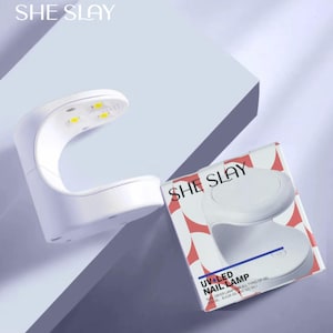 May include: A white UV+LED nail lamp with the text "SHE SLAY" on the top and on the box. The box also has the text "UV+LED NAIL LAMP". The lamp has a curved design with four yellow lights.