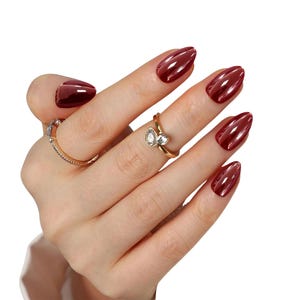 May include: A hand with polished, maroon-colored fingernails. The nails are oval-shaped and have a glossy finish. Two gold rings with clear stones adorn the fingers. The background is plain white.