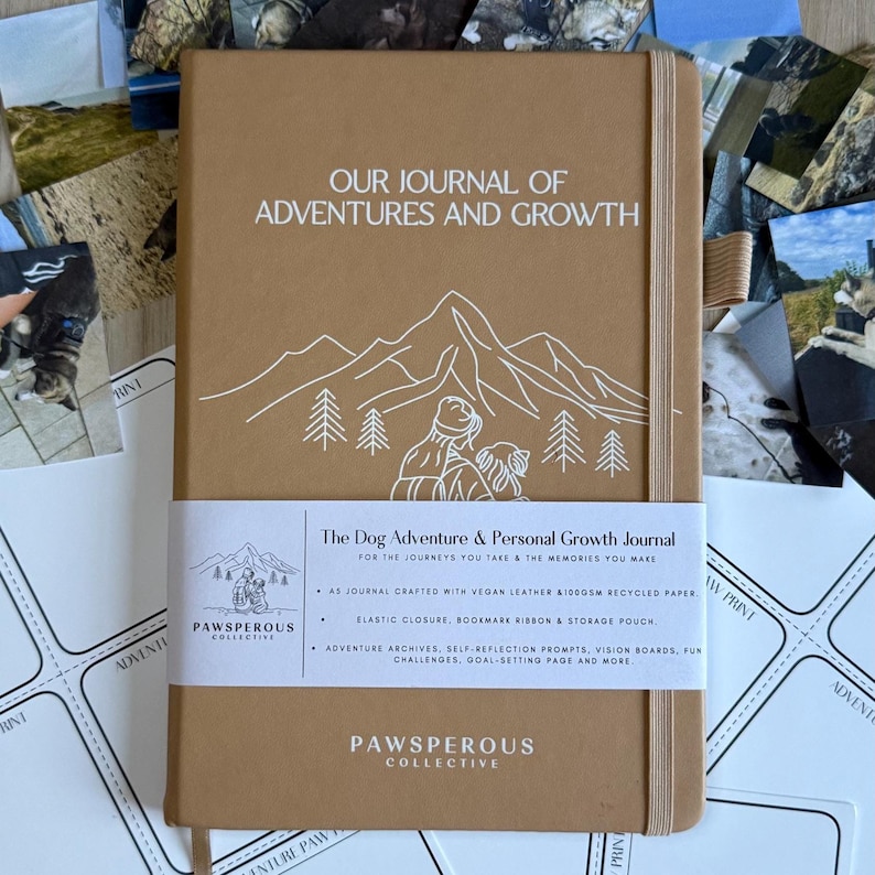 Op de afbeelding: Een bruin notitieboek met de tekst "OUR JOURNAL OF ADVENTURES AND GROWTH" en een illustratie van een berg en een hond. Het notitieboek heeft een witte band met de tekst "The Dog Adventure & Personal Growth Journal" en het logo "PAWSPEROUS COLLECTIVE".