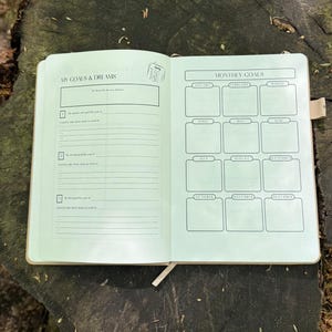Op de afbeelding: Open notitieboek met de titels "MY GOALS & DREAMS" en "MONTHLY GOALS". De linkerpagina heeft prompts voor jaarlijkse doelen. De rechterpagina heeft vakken voor elke maand, van januari tot december. Het notitieboek ligt open op een houten oppervlak.