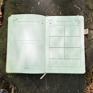 Op de afbeelding: Een open dagboek met lichtgroene pagina's, ontworpen voor vision boards. De linkerpagina is voor avonturen, de rechter voor levensdoelen. Secties zijn onder meer carri&egrave;re, familie en reizen. Het dagboek ligt open op een houten oppervlak, met een bladwijzer.