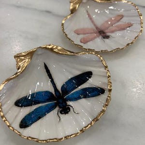 May include: Two decorative seashell dishes. One features a blue dragonfly, the other a pink dragonfly. Both have gold-painted rims and are set against a white background.
