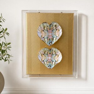 May include: A rectangular clear acrylic shadow box frame containing a gold-colored textured background. Two heart-shaped decorative objects with a multi-colored, marbled, and speckled pattern in shades of pink, blue, and green are mounted vertically.