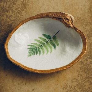 May include: A decorative seashell bowl with a gold-painted rim and a white interior. A green fern leaf design is painted on the inside of the bowl. The bowl is set against a brown background with a floral pattern.