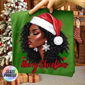 May include: Green t-shirt featuring a graphic of a woman wearing a Santa hat and snowflake earrings. The text "Merry Christmas" is written in red script. The woman has curly black hair and festive makeup.