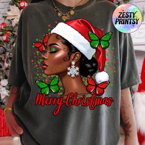 May include: Dark gray t-shirt featuring a graphic of a woman wearing a Santa hat and snowflake earrings, with green and red butterflies. The words "Merry Christmas" are written in red script.