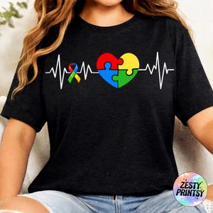 以下が含まれることがあります： 自閉症啓発デザインの黒いTシャツ。マルチカラーのリボン、ハート型のパズル、白い心拍ラインが含まれています。シャツの右下には「Zesty Prints」の文字が入っています。