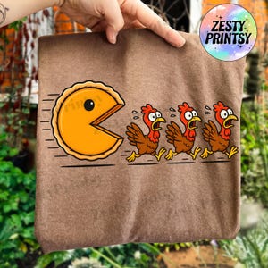May include: A brown t-shirt featuring a cartoon pie character chasing three frightened chickens. The pie is yellow with a black eye and a bite taken out of it. The chickens are brown with red combs and are running away.