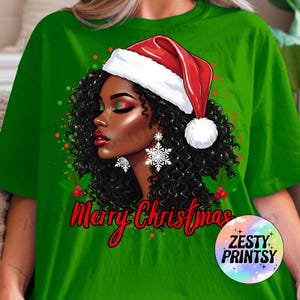 May include: Green t-shirt featuring a graphic of a woman with curly black hair wearing a Santa hat and snowflake earrings. The words "Merry Christmas" are written in red script. A circular logo with the words "Zesty Prints" is in the lower right corner.