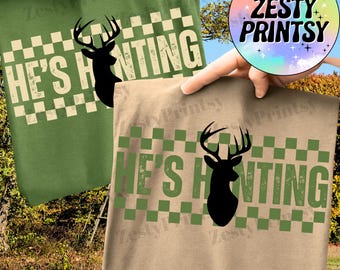 May include: Two t-shirts, one green and one tan, each with a deer silhouette and the text "HE'S HUNTING" in a green and white checkered design. The shirts are held up against a natural outdoor background.