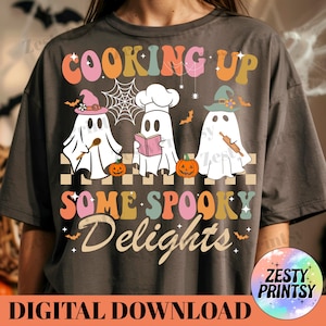 May include: Dark gray t-shirt with a colorful graphic that reads "COOKING UP SOME SPOOKY DELIGHTS." The design features three ghost characters wearing chef hats and witch hats, with pumpkins and bats. The words "DIGITAL DOWNLOAD" are at the bottom.
